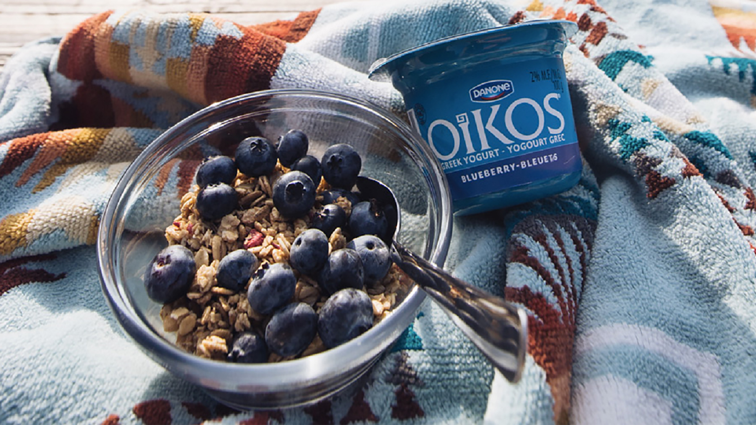 Un yaourt Oikos blueberry et bleuets à côté d'un bol de céréales et de myrtilles